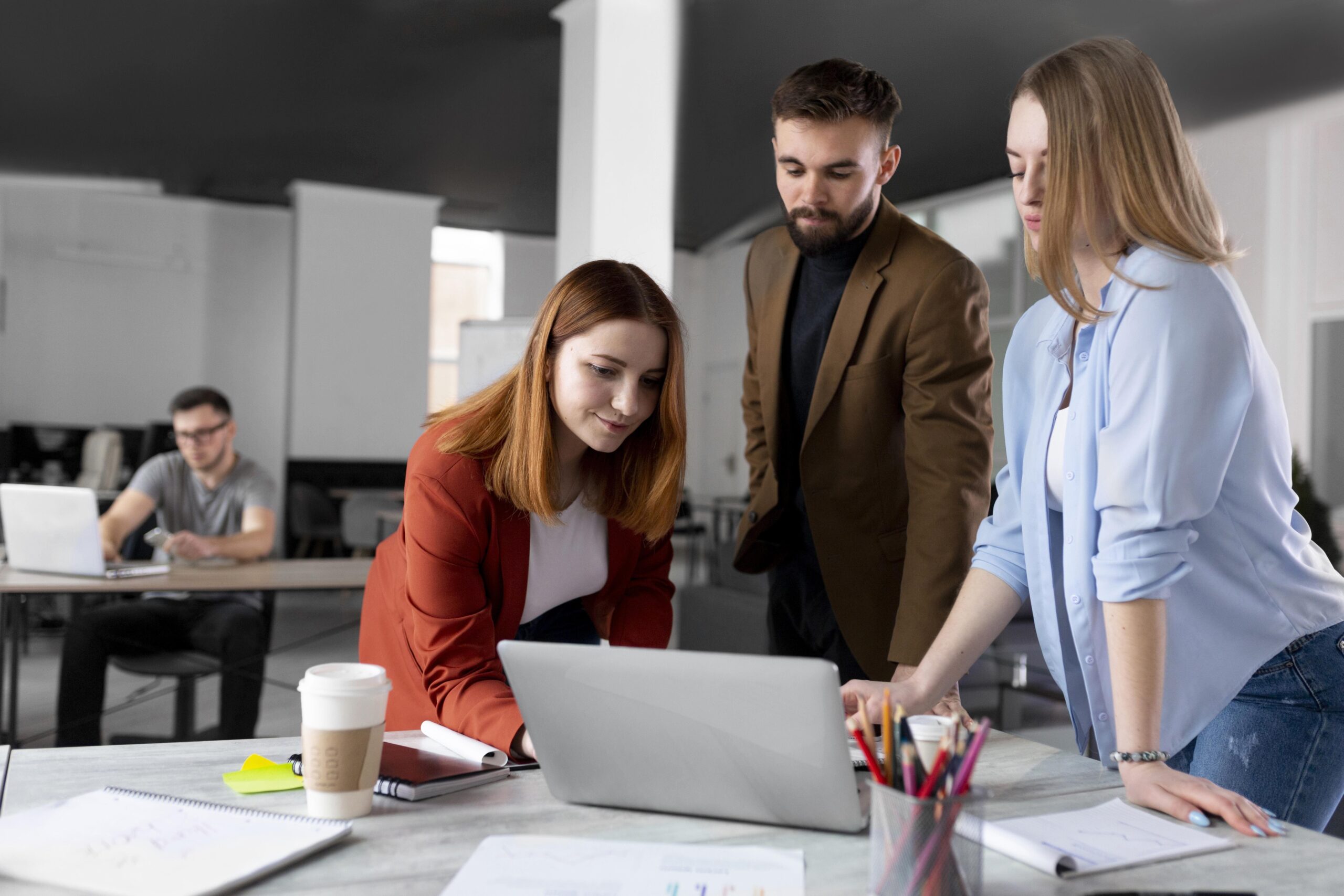 Un grupo de personas trabajando en equipo alrededor de una mesa, concentrados en la pantalla de una computadora portátil. En el fondo, otra persona trabaja individualmente en un escritorio.