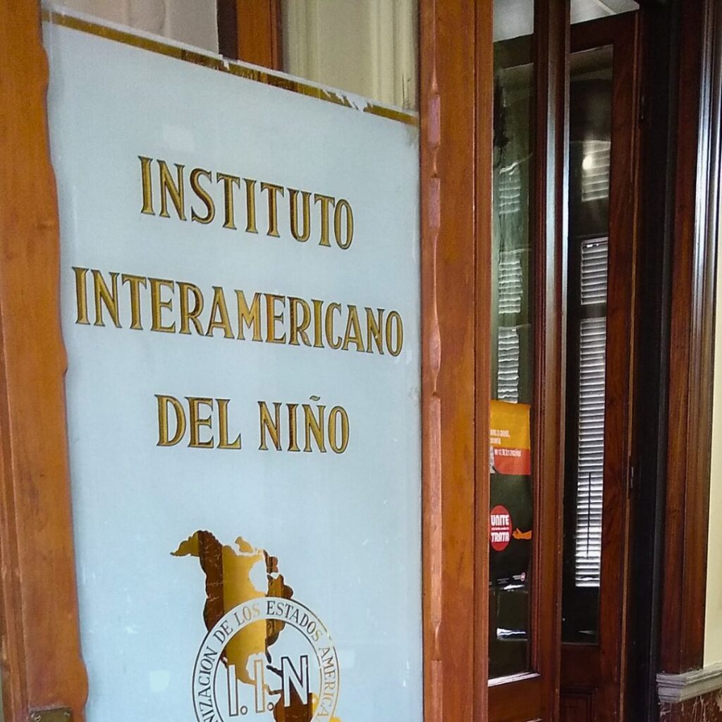 Wooden door with the text 'Instituto Interamericano del Niño' in gold letters and the emblem of the Organization of American States (OAS). The door opens into a classic interior with a marble floor, tiled patterns, and a wooden staircase