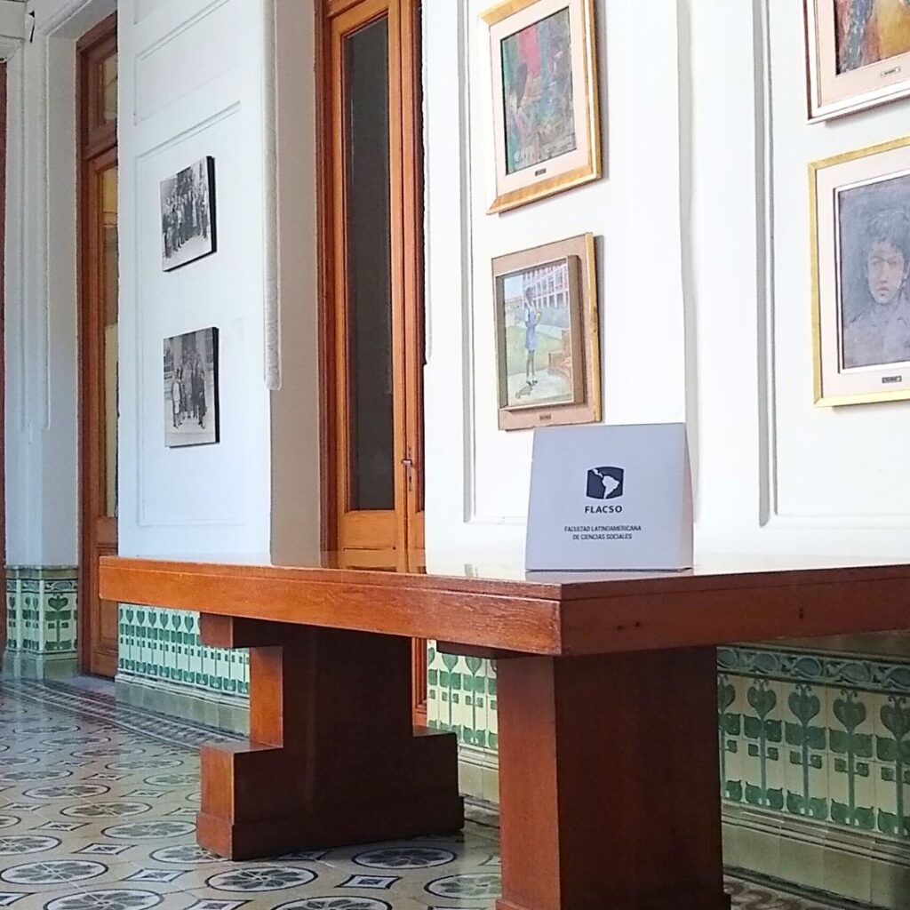 A brightly lit hallway with decorative patterned floor tiles and green tiled walls. The space features wooden doors, framed paintings on the walls, and a wooden bench with a sign reading 'FLACSO' on top