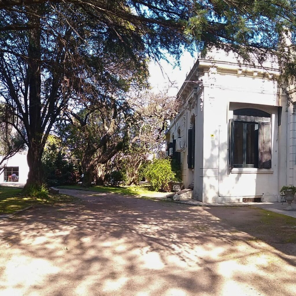 A sun-dappled view of a large, white, Spanish-style house partially obscured by the shadows of overhanging trees.  The house features dark window shutters and is set back from a dirt path. Lush greenery and mature trees surround the property